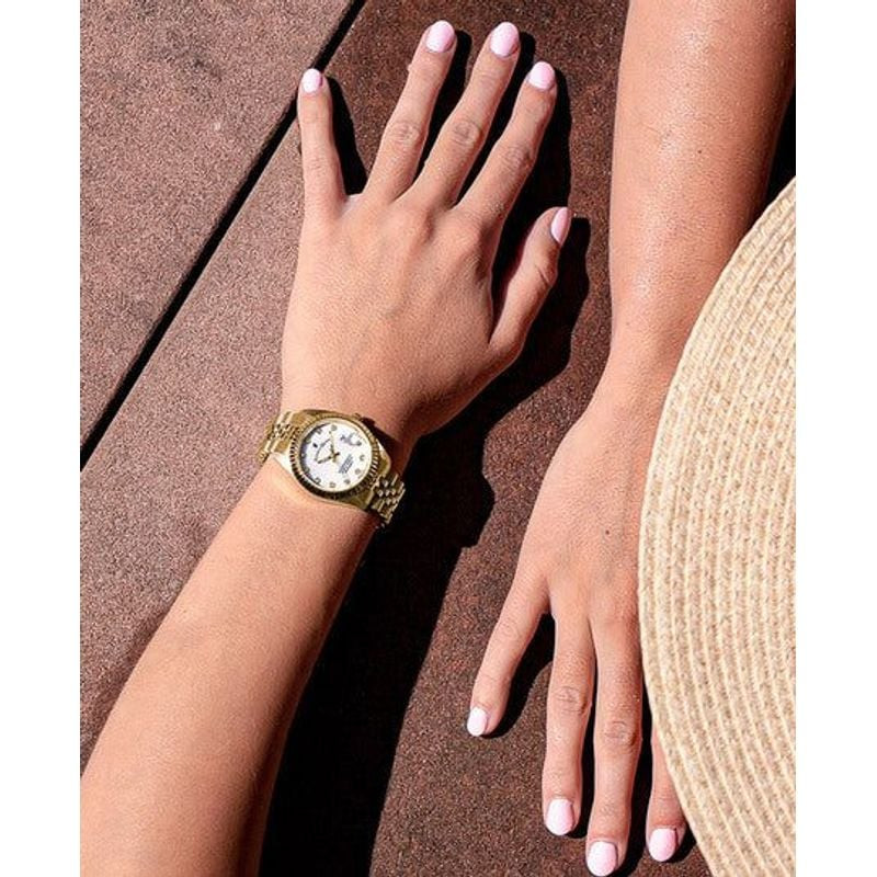 Poignet d'une femme portant la montre Jacques du Manoir Inspiration dorée en extérieur près d'un cha
