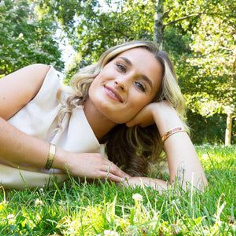 Femme blonde allongée sur l'herbe verte dans un jardin, portant le bracelet jonc marguerite doré Lou