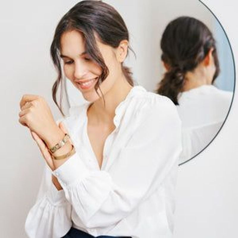 Femme en chemise blanche souriante portant le bracelet manchette Monstera doré au poignet, miroir de