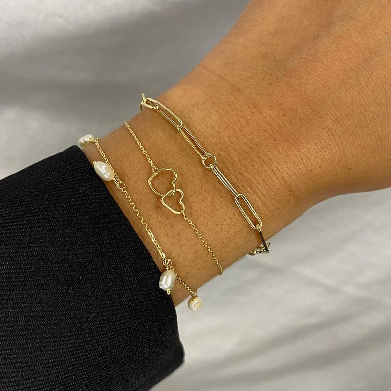 Bracelet Double Cœur en or 14 carats porté au poignet, superposé à des bracelets de perles et chaîne