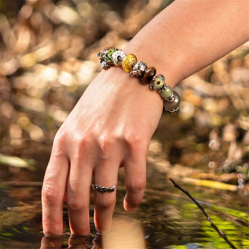 Main portant un bracelet Trollbeads, doigts plongés dans un ruisseau forestier, perles en verre et c