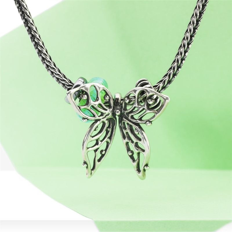 Perle Plume turquoise Trollbeads sur une chaîne en argent derrière un pendentif papillon sur fond ve