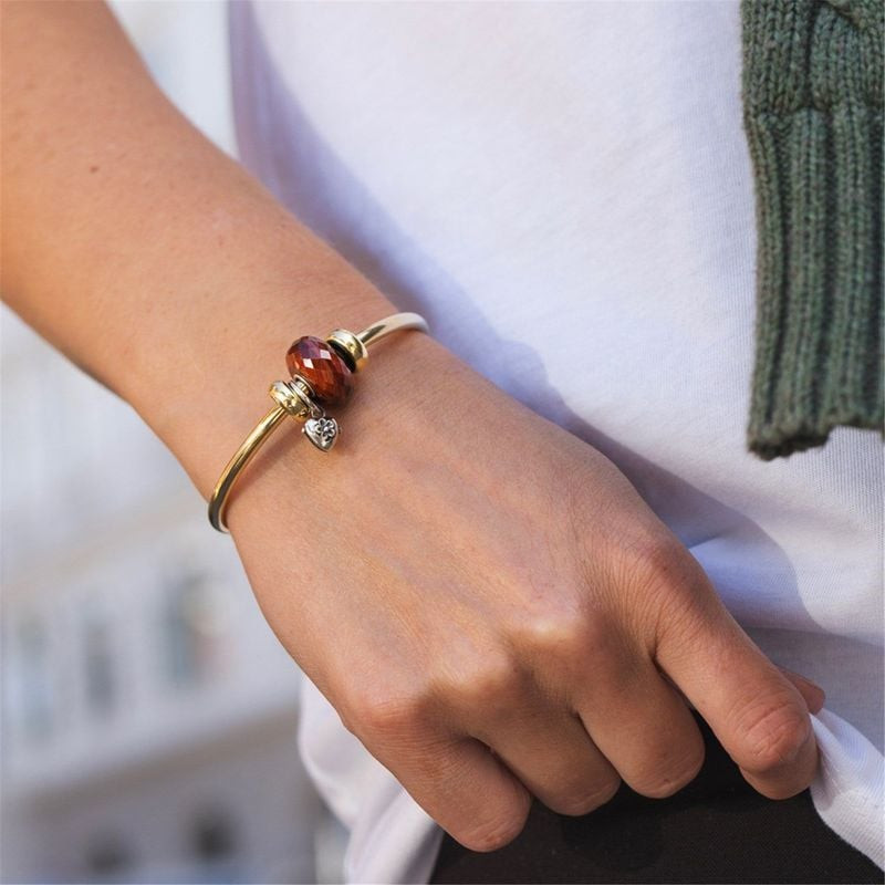 Poignet portant un jonc doré avec la perle Trollbeads œil de tigre rouge, main posée sur une chemise