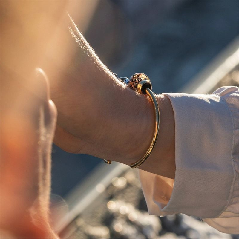 Poignet portant un jonc Trollbeads doré avec la perle Happy 2023 en verre noir et doré sous le solei