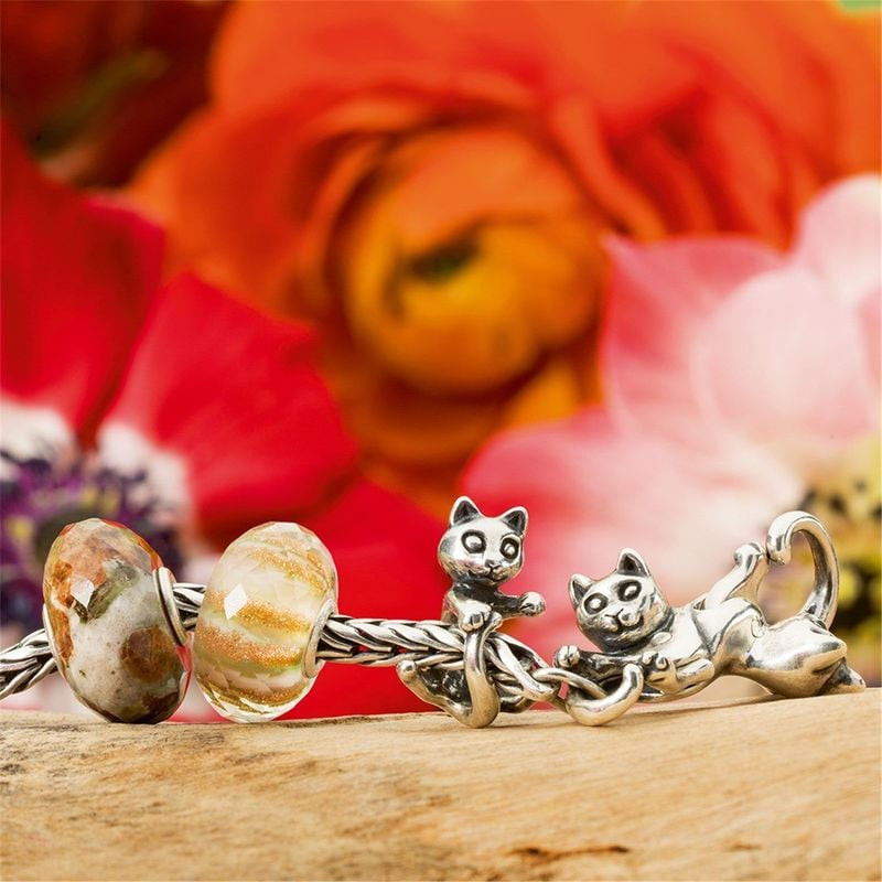 Un bracelet Trollbeads orné de la perle Lit de Fleurs et d'une breloque chat espiègle, sur un fond f