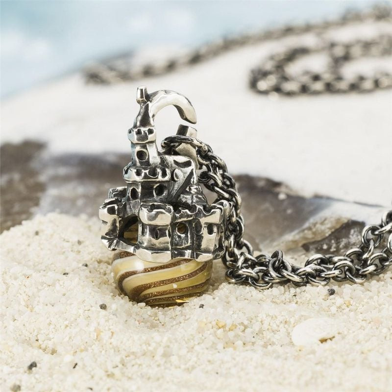 Pendentif Trollbeads Château de sable posé sur le sable blanc avec un coquillage et l'océan flou en 